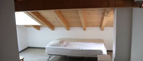 Pequeña habitación con cama individual, techo inclinado y vigas de madera, una mesita y una escalera.