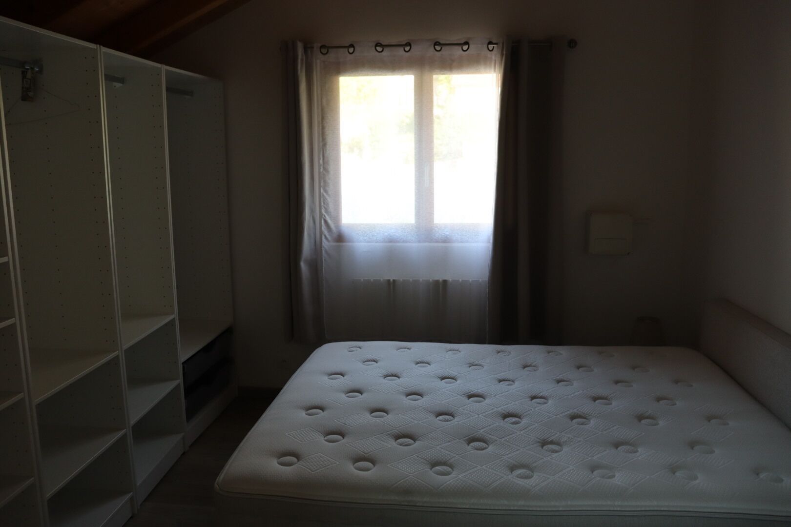 Une pièce avec un seul matelas, une armoire et une fenêtre avec des rideaux.