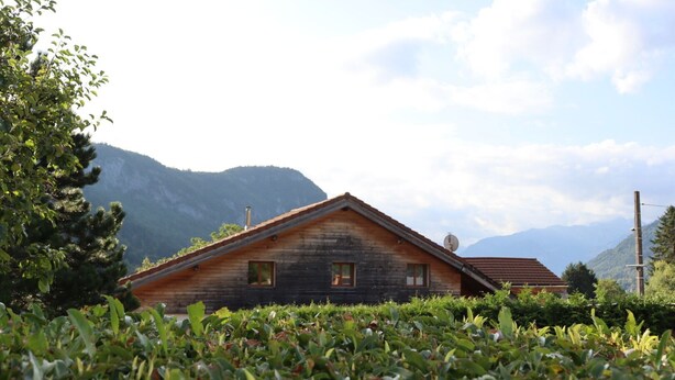 A wooden cabin with a sloped roof, surrounded by lush greenery and mountains in the background.
