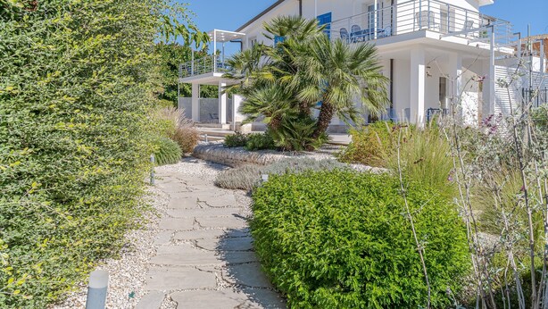 A modern two-story house with a balcony, a stone pathway, and lush greenery.