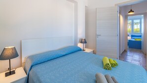 A bedroom with a bed, two bedside lamps, a door leading to another room, and a view of a balcony.