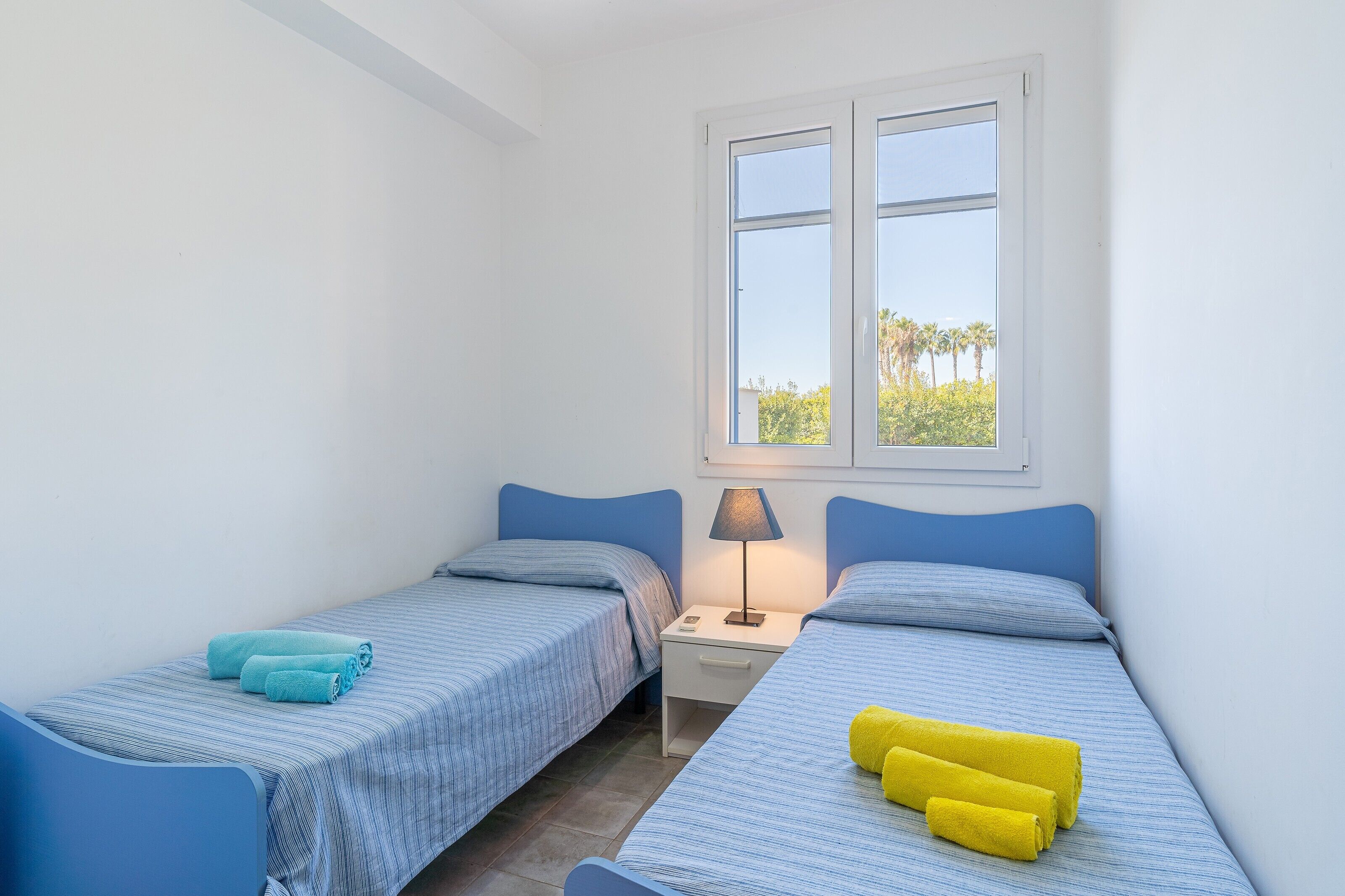 Une chambre avec deux lits simples, une table de chevet avec une lampe et une fenêtre donnant sur des palmiers.