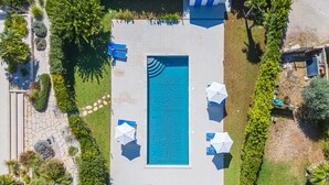 An aerial view of a rectangular swimming pool surrounded by a stone pathway, with blue umbrellas and lounge chairs.