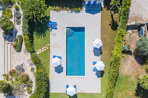 Une vue aĂ©rienne dâune piscine rectangulaire entourĂ©e dâun chemin en pierre, avec des parasols bleus et des chaises longues.