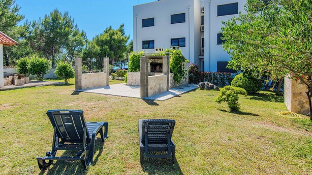 An outdoor seating area with two chairs, a stone bench, and a small fire pit in front of a modern building with multiple windows. - Holiday home Kurta  (Privlaka)