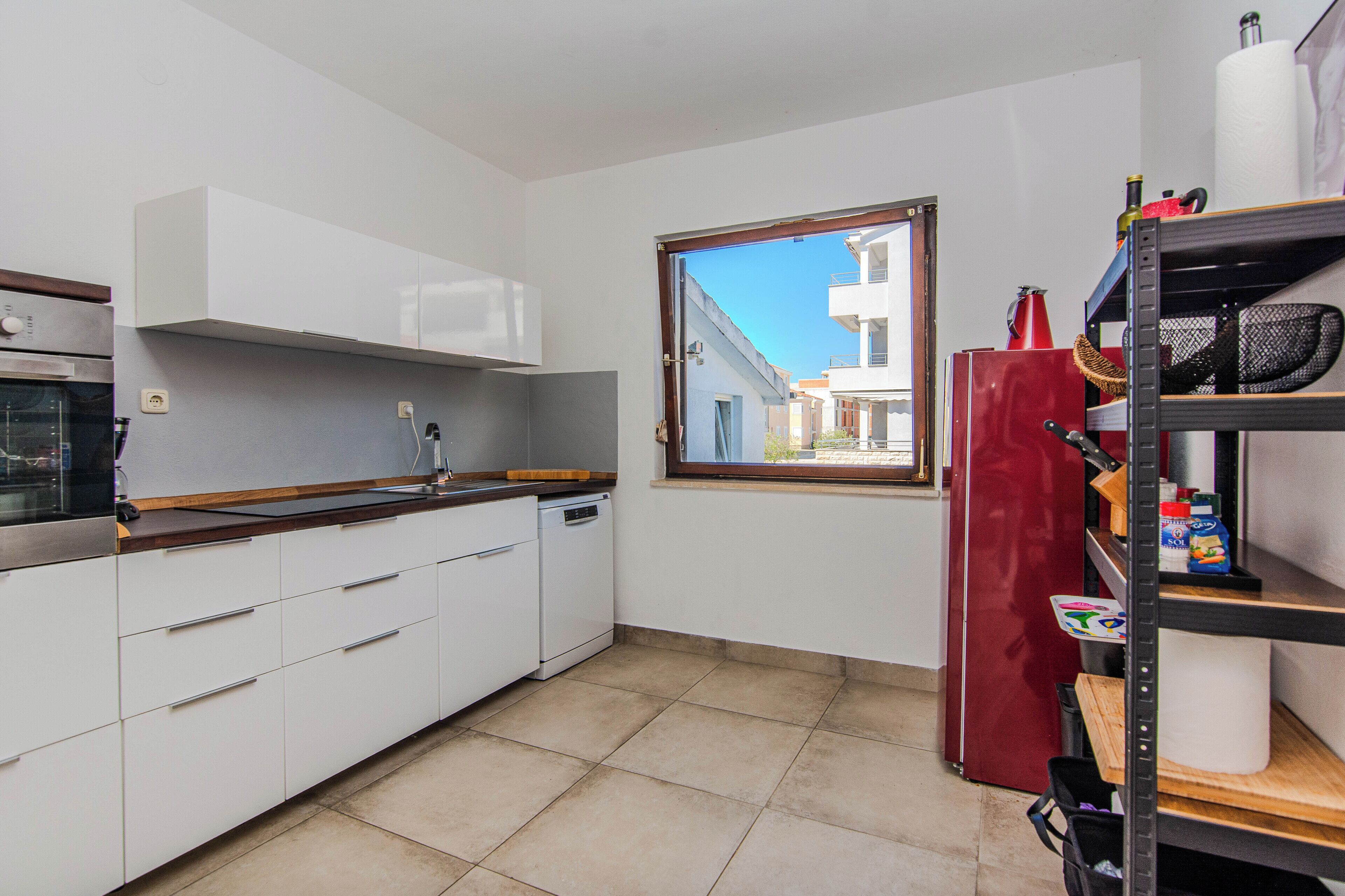 Une cuisine de taille modeste, avec des meubles blancs, un réfrigérateur rouge et une fenêtre donnant sur un quartier résidentiel.