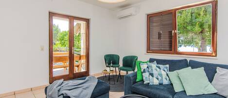 Una sala de estar con un sofá azul, dos sillones verdes, una mesa de centro y un amplio ventanal con persianas.