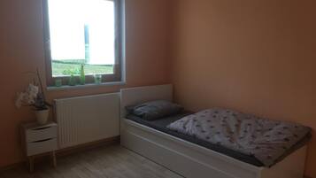 A bedroom with a single bed, a radiator, a window with a view of greenery, and a small bedside table with a plant.