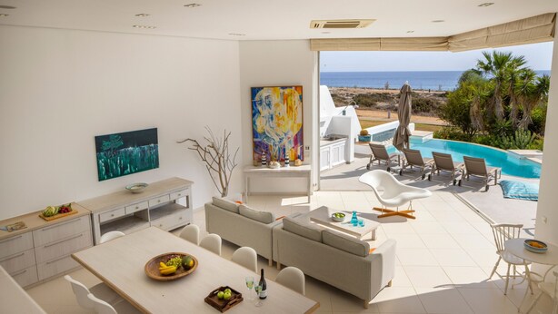 A modern living room with a dining area, a large painting, and a view of the beach and pool. - Protaras Seafront Villa Alemar (Paralimni)