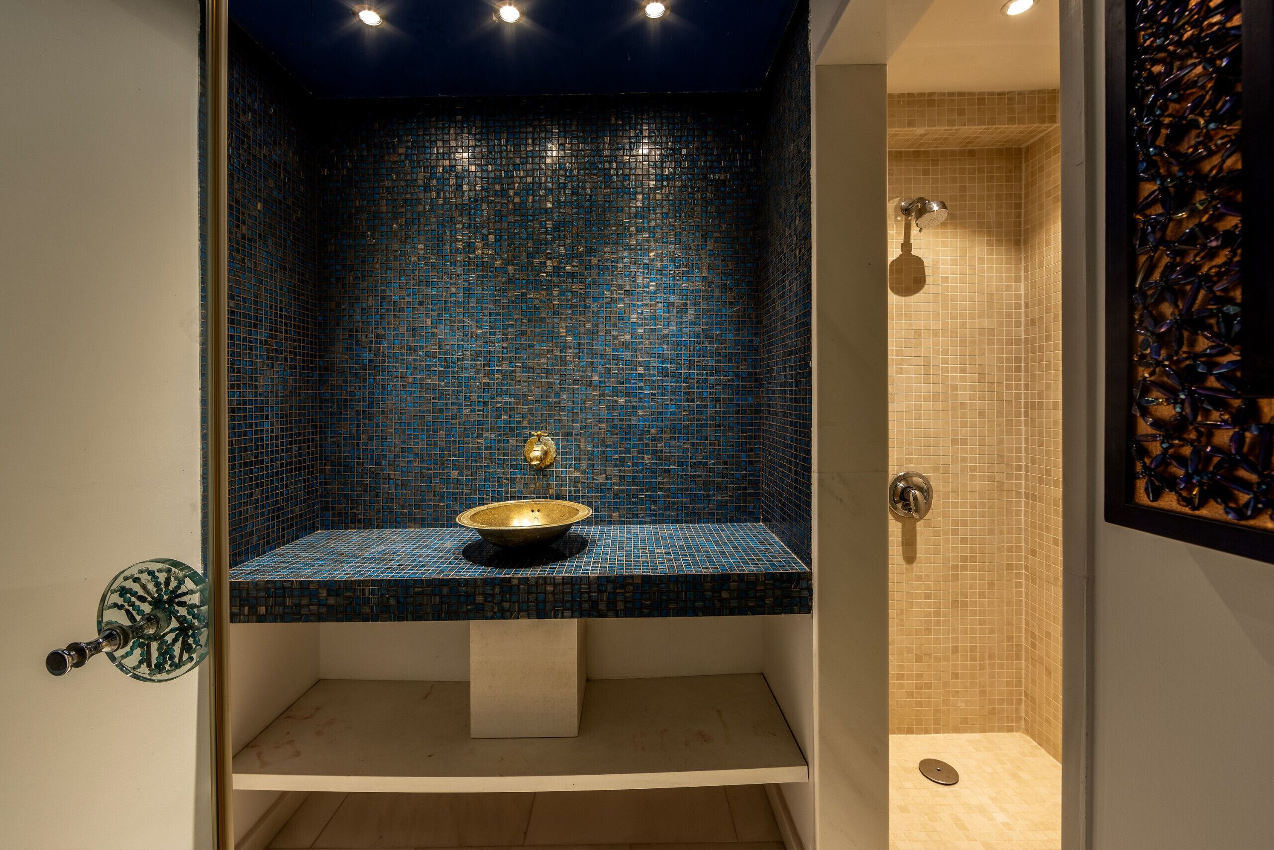Un baño con pared de mosaico azul, lavamanos dorado y zona de ducha.