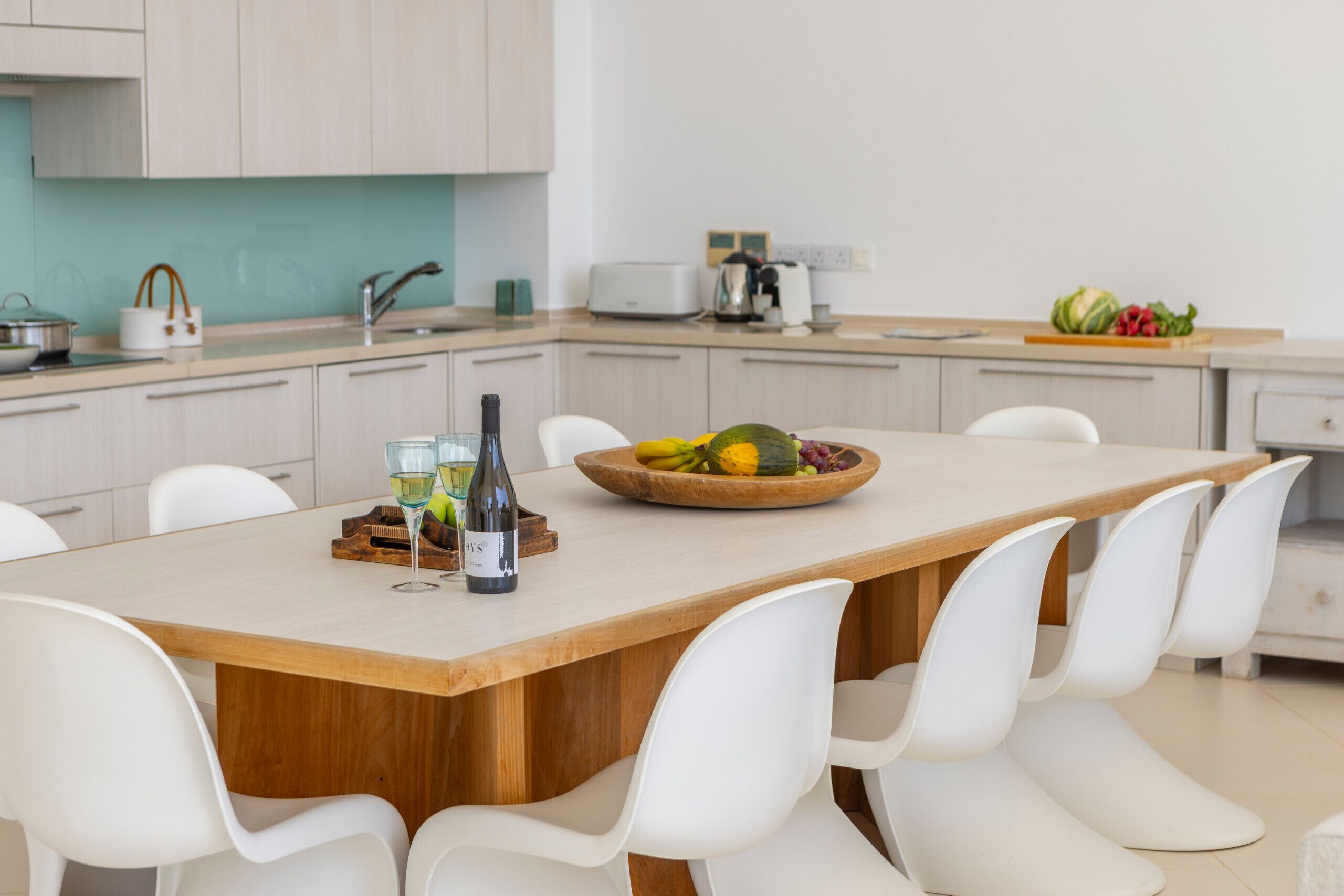 A modern kitchen with a central island, white chairs, a fruit bowl, and a wine bottle.