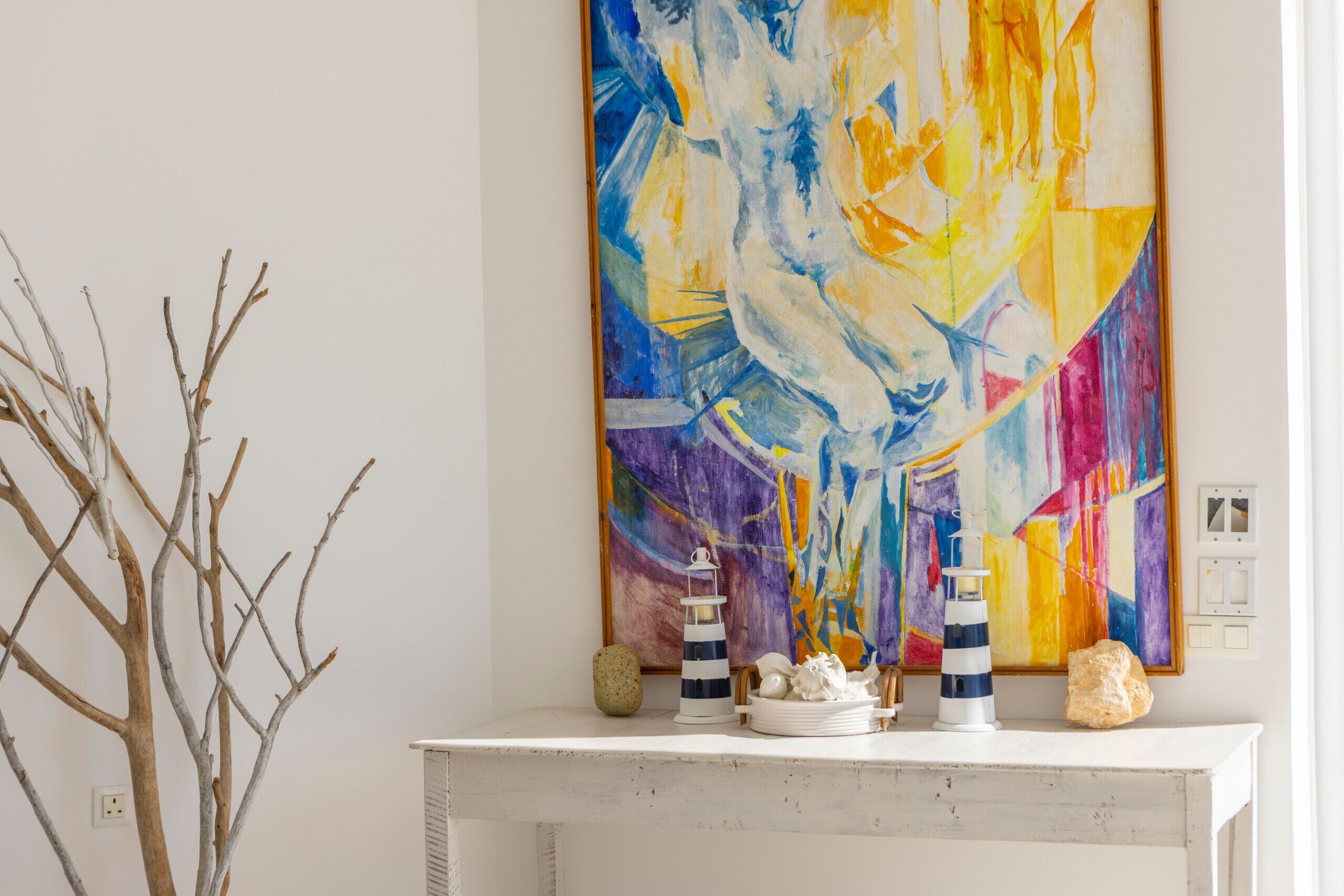Une console blanche, ornée d’un tableau abstrait aux couleurs vives, d’une décoration représentant un phare et d’une petite branche d’arbre.