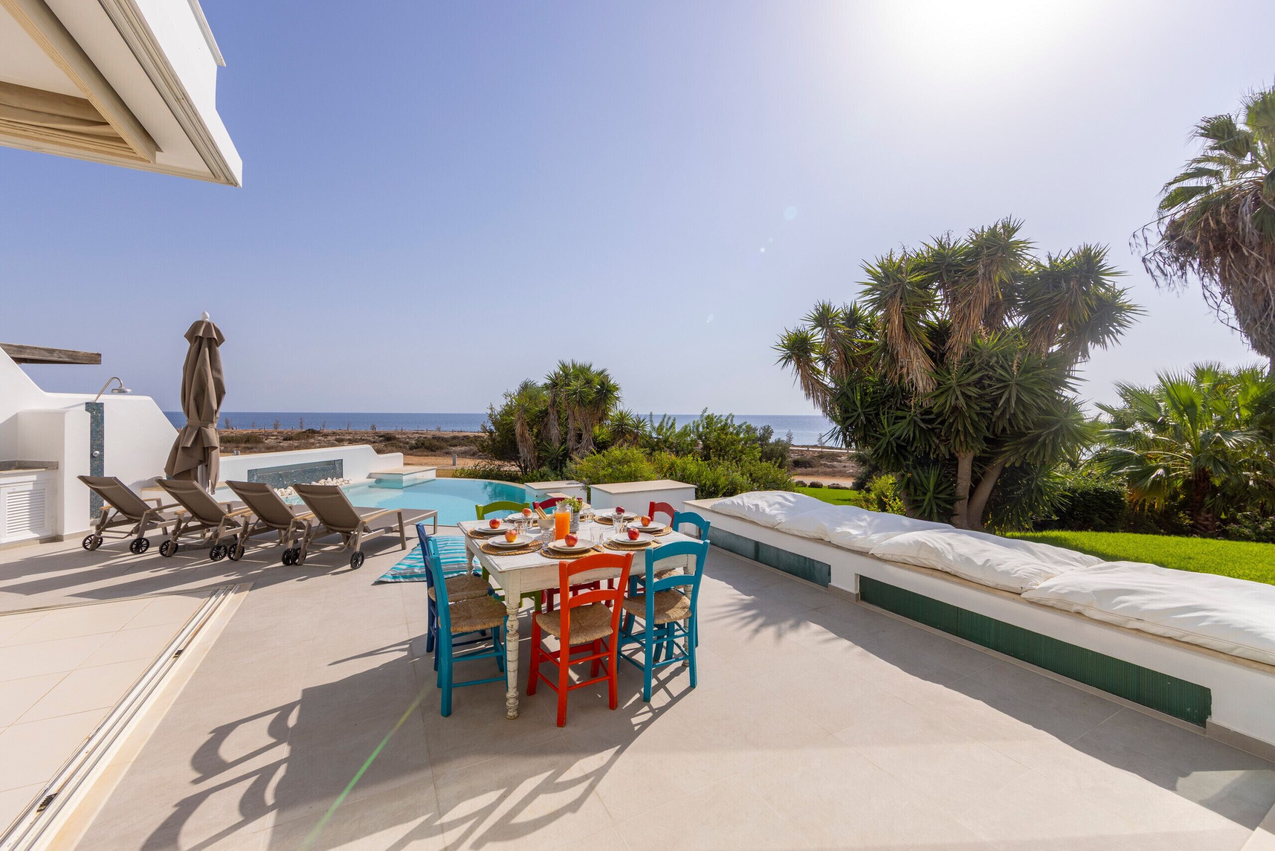 A poolside area with a table and chairs, a pool, and a view of the sea.