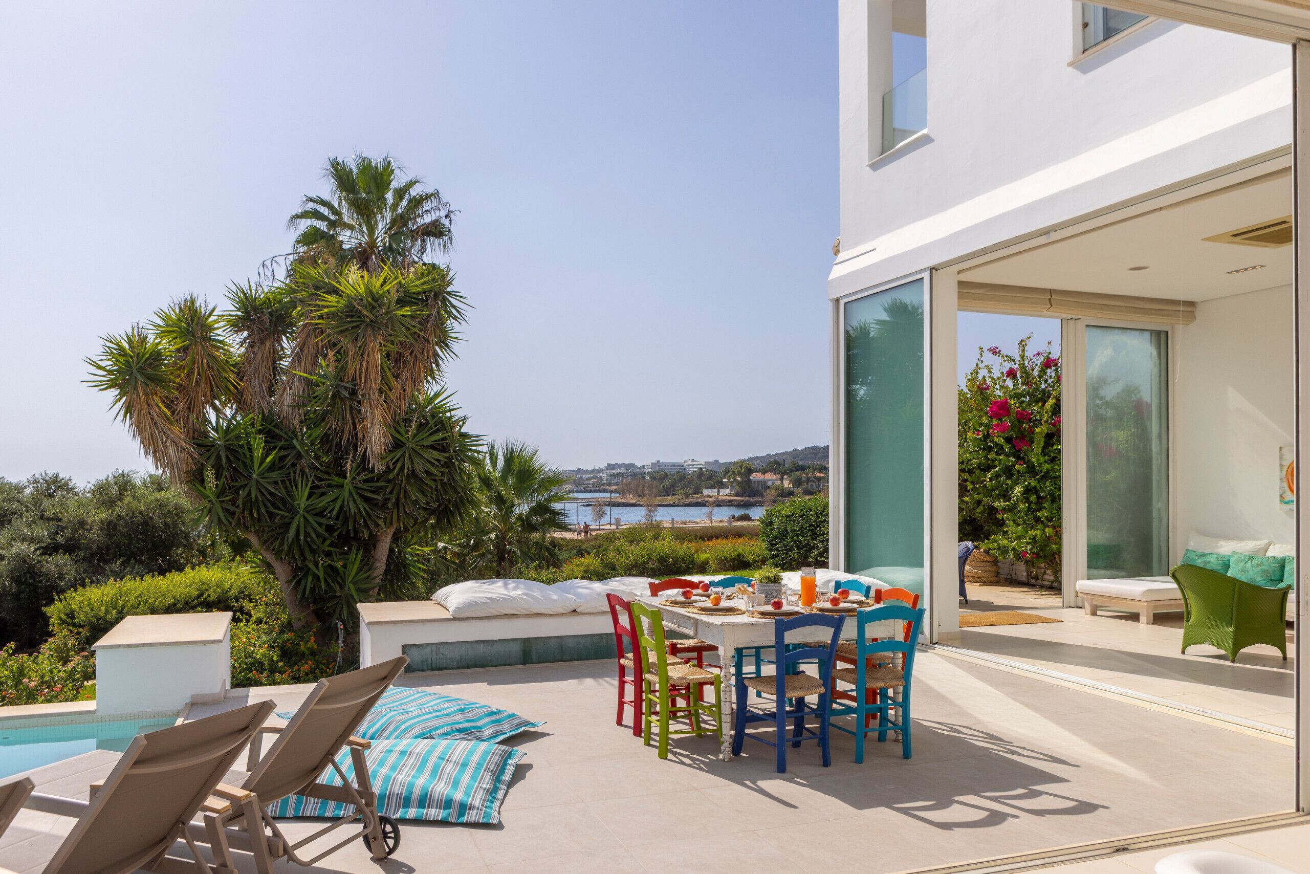 A poolside area with a table and chairs, a lounge chair, and a view of the sea.