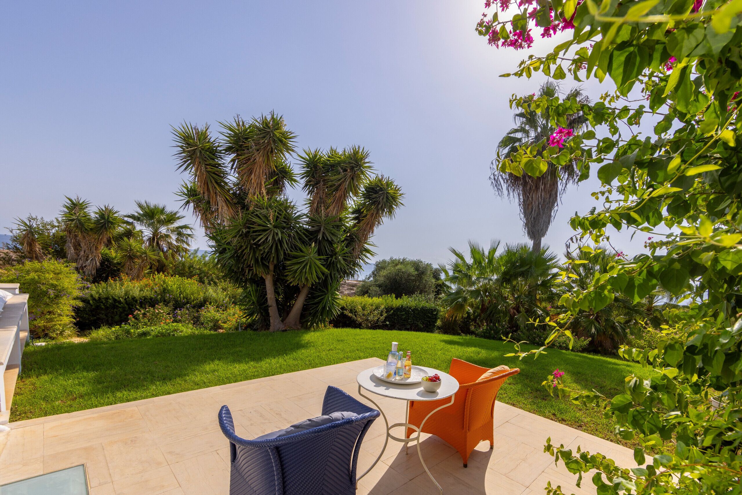 A patio with a round table and two chairs, surrounded by lush greenery and palm trees.