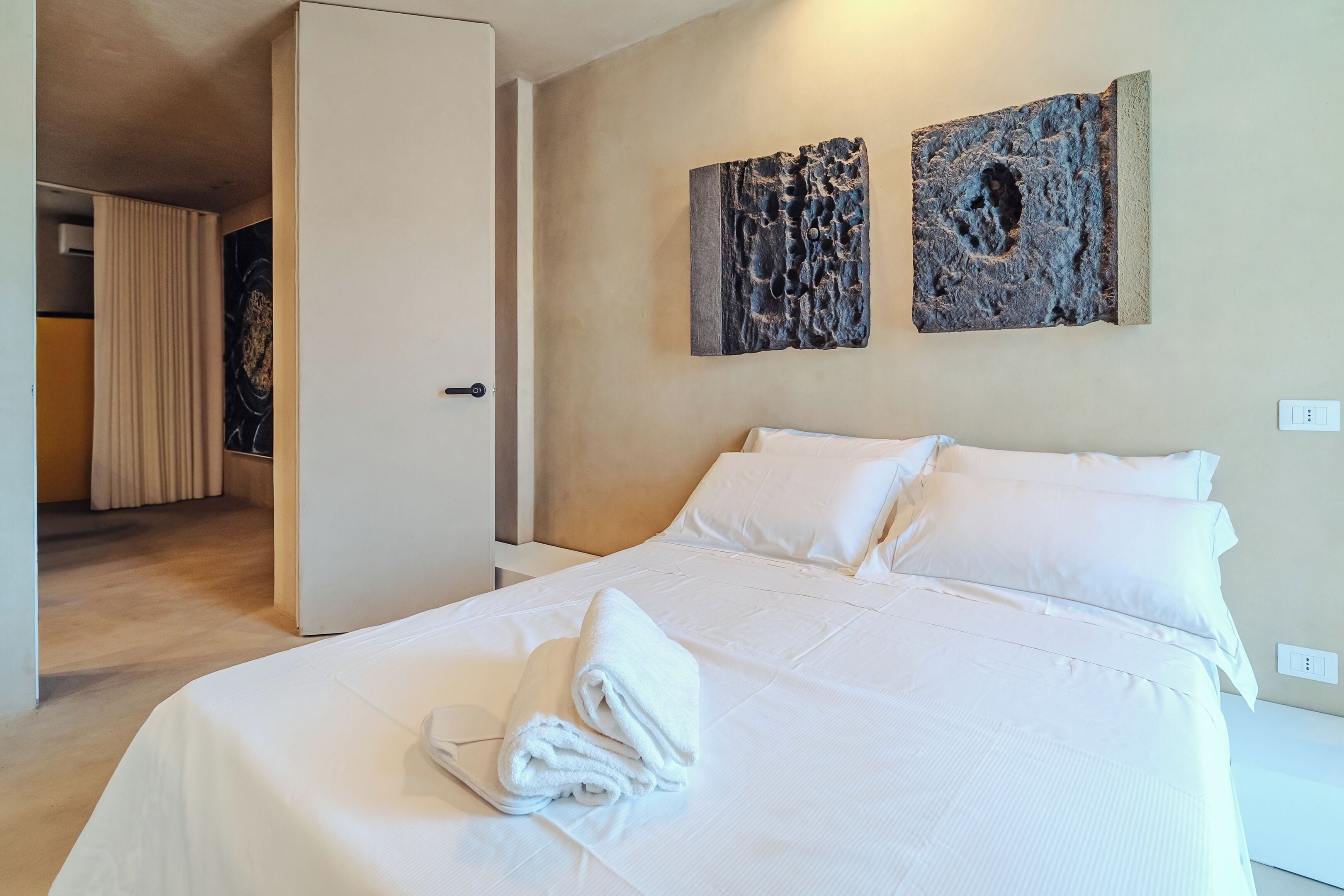 A hotel room with a neatly made bed, a door leading to another room, and two abstract wall art pieces.