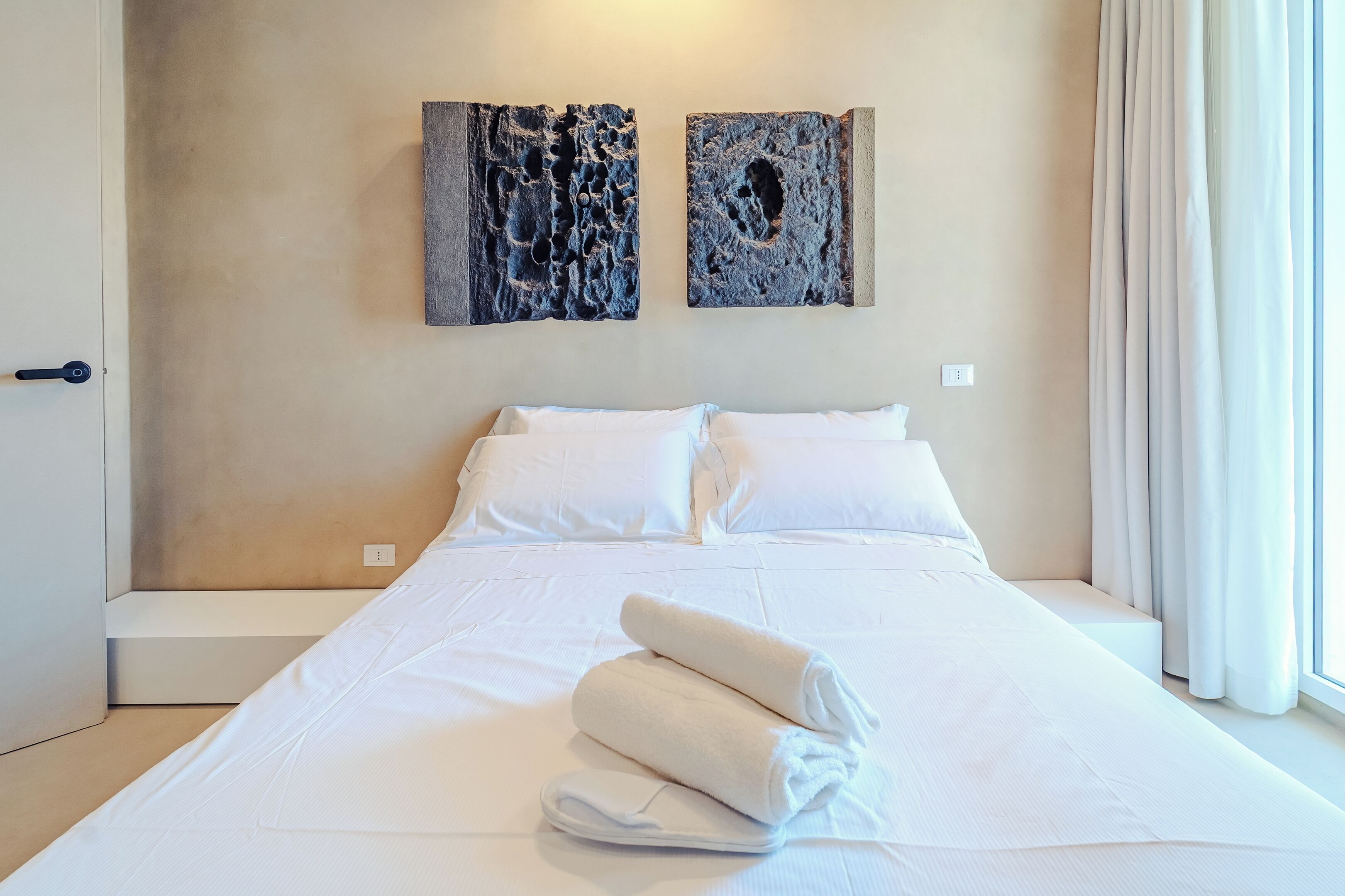 Habitación de hotel con una cama grande, ropa de cama blanca, dos obras de arte abstractas en la pared y una ventana con cortinas.