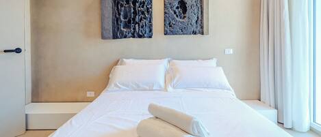 Habitación de hotel con una cama grande, ropa de cama blanca, dos obras de arte abstractas en la pared y una ventana con cortinas.