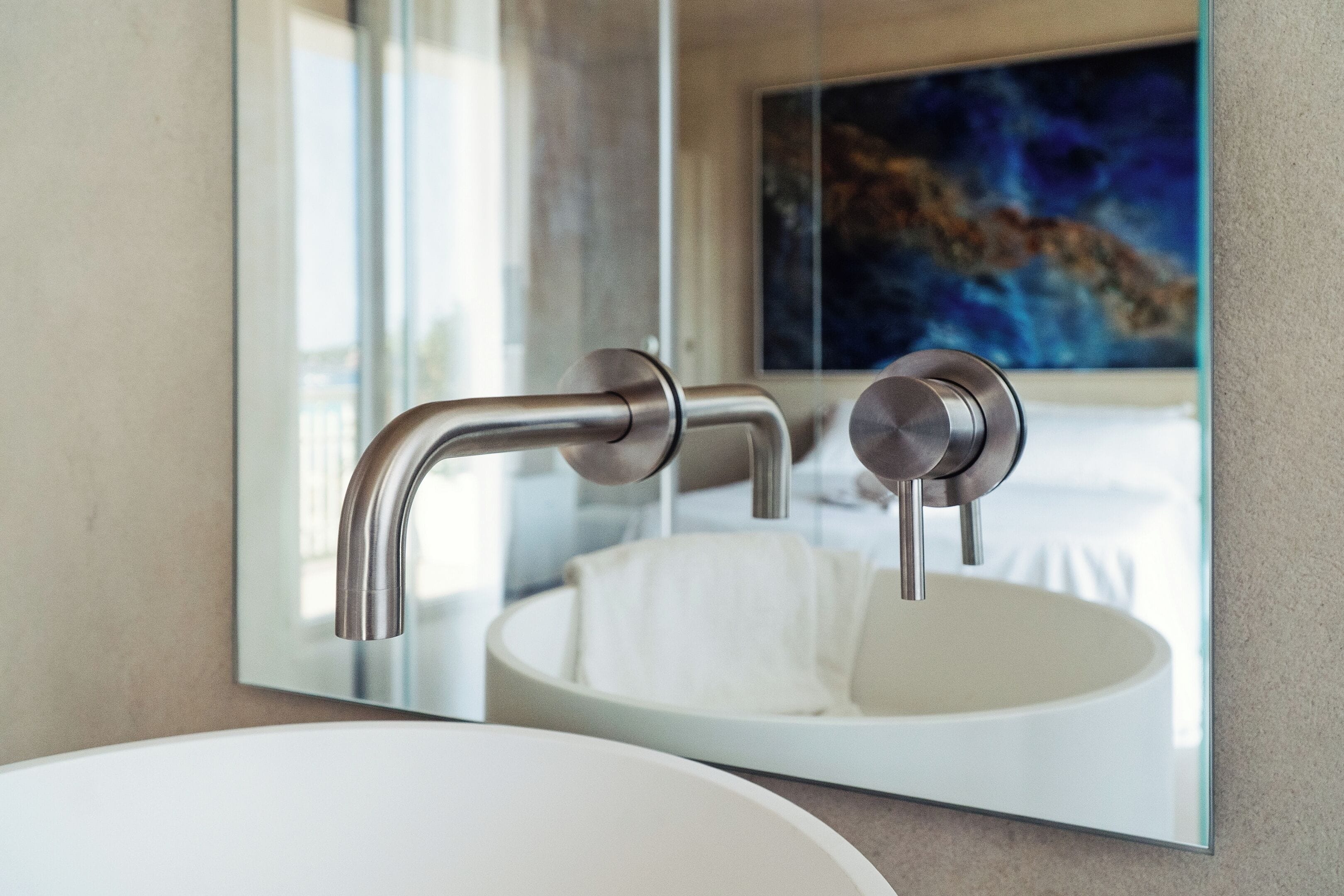 Een moderne badkamer met een witte wastafel, een strakke kraan en een grote spiegel die een bed en een schilderij weerspiegelt.