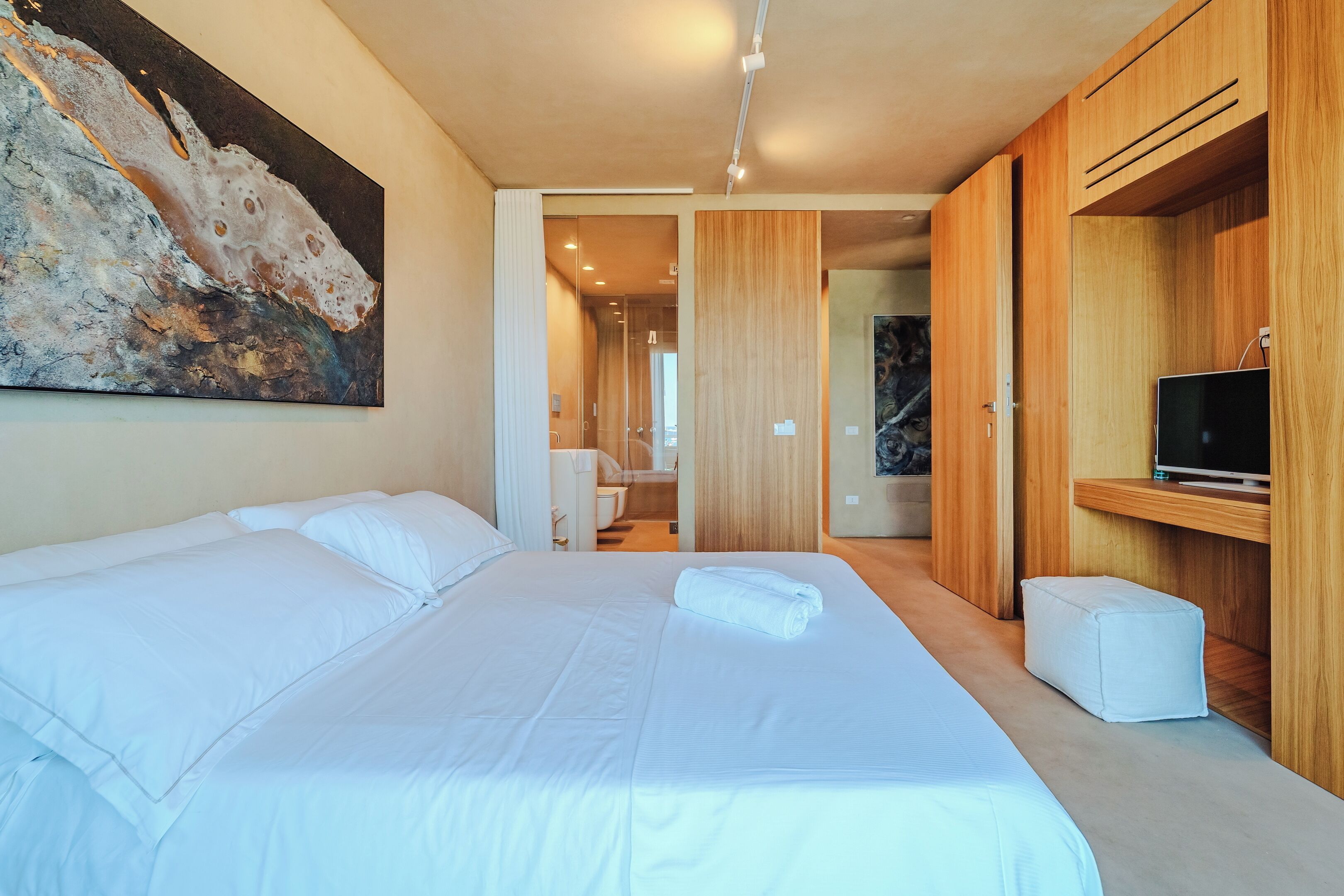A modern hotel room with a large bed, a painting on the wall, wooden cabinets, and a television.