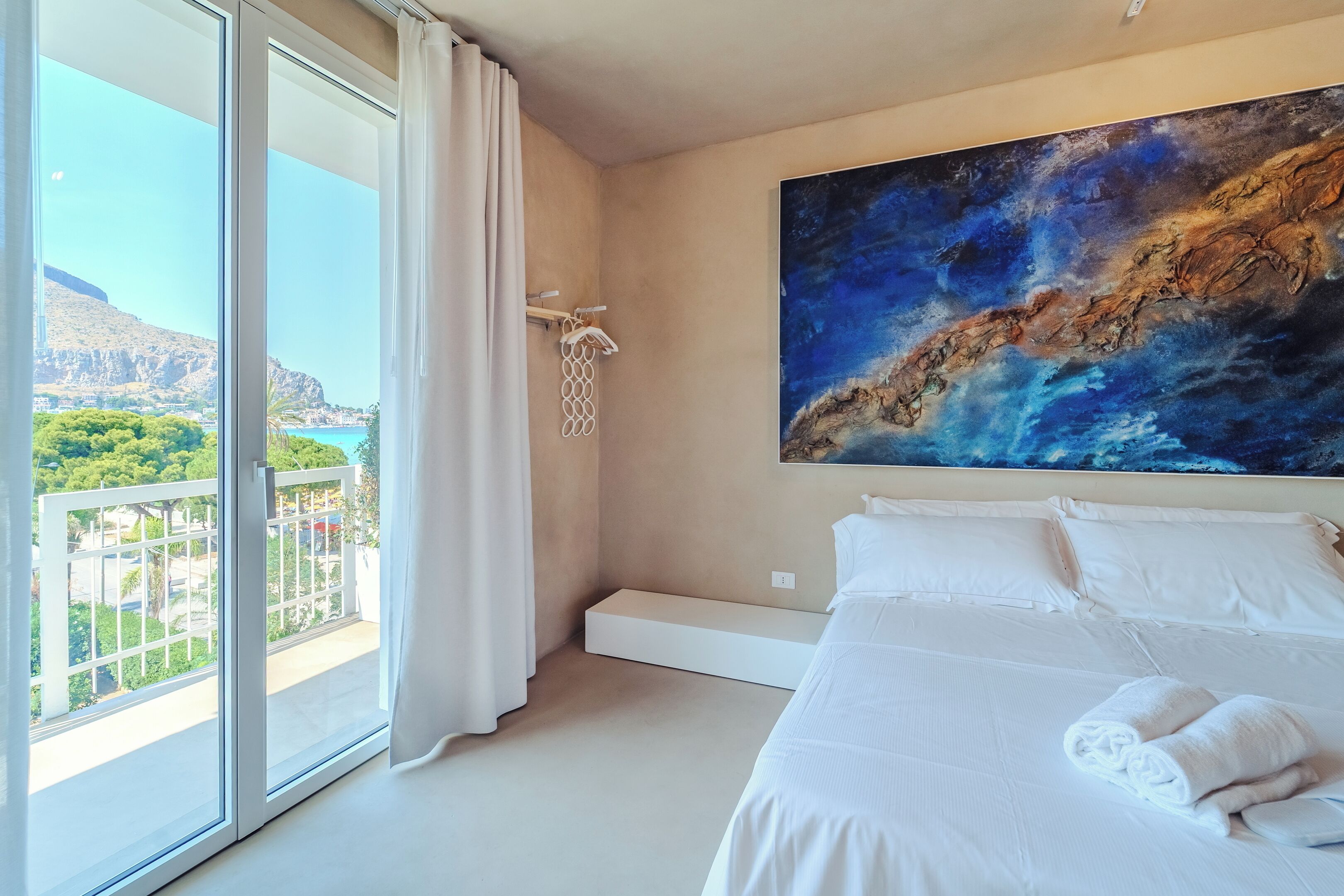 Ein Hotelzimmer mit einem großen Bett, weißer Bettwäsche, einem Bild an der Wand und einem Balkon mit Blick auf Grünflächen und ein Gebirge.