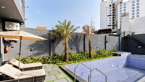 A rooftop pool with lounge chairs, a small garden with palm trees, and a view of a cityscape.