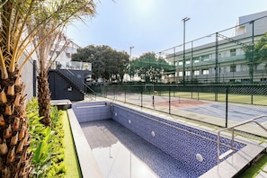 Piscina com design de mosaico azul, cercada por uma grade de metal e adjacente a um edifício com fachada de vidro.