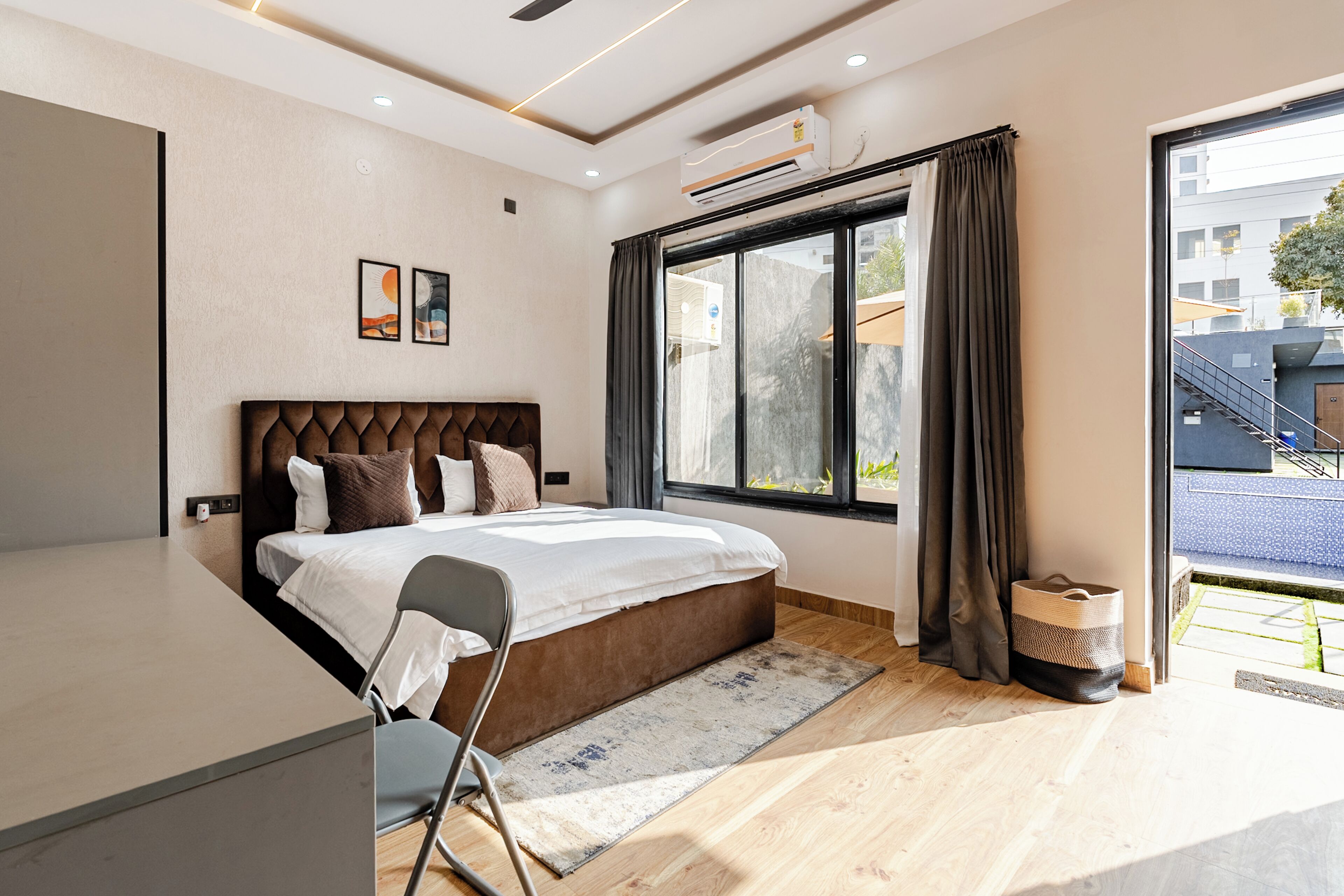 A modern bedroom with a bed, a desk, a chair, a large window with curtains, and an air conditioning unit.