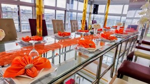 Una larga mesa con tapas de cristal y barandillas de metal, decorada con cintas naranjas y flores, en una habitación moderna con ventanas amplias y vistas a la ciudad.