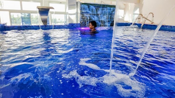 Una piscina interior con una persona nadando, una cascada y vistas de la ciudad a través de amplias ventanas.