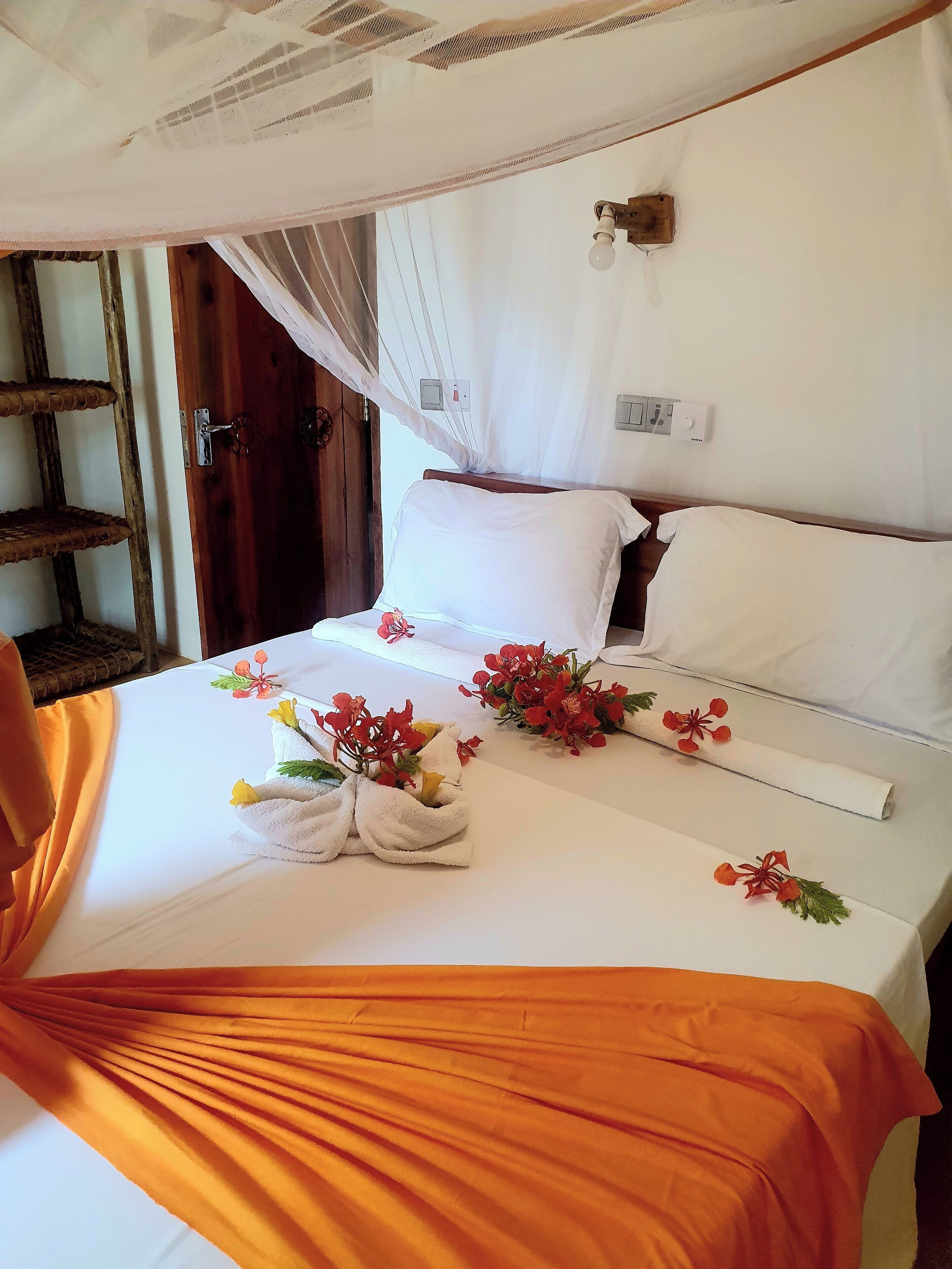 Una cama bien hecha con sábanas blancas, cabecera de madera y dosel. Hay toallas decorativas con flores y un cortinaje naranja.