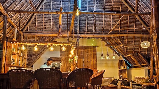A rustic interior with a high ceiling, wooden beams, and a thatched roof. There are wicker chairs and a wooden table in the foreground, and a bar area in the background. - SuBanda Boutique Hotel (Makunduchi)