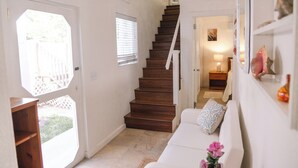 A staircase leading to a second floor, a white sofa with a cushion, a wooden shelf, and a window with blinds. - Charming Tickled Pink 2BR, 3BD, 3BTH home, minutes from pink sand beach (Harbour Island)