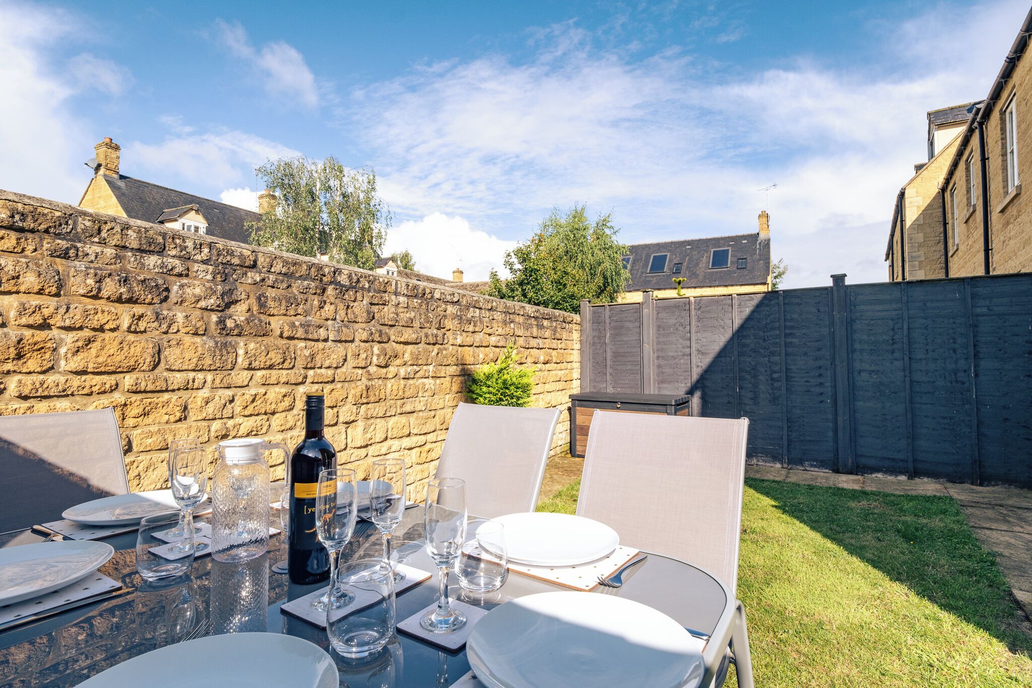 A table set outdoors with wine glasses, plates, and a bottle, surrounded by chairs, against a stone wall and a wooden fence.