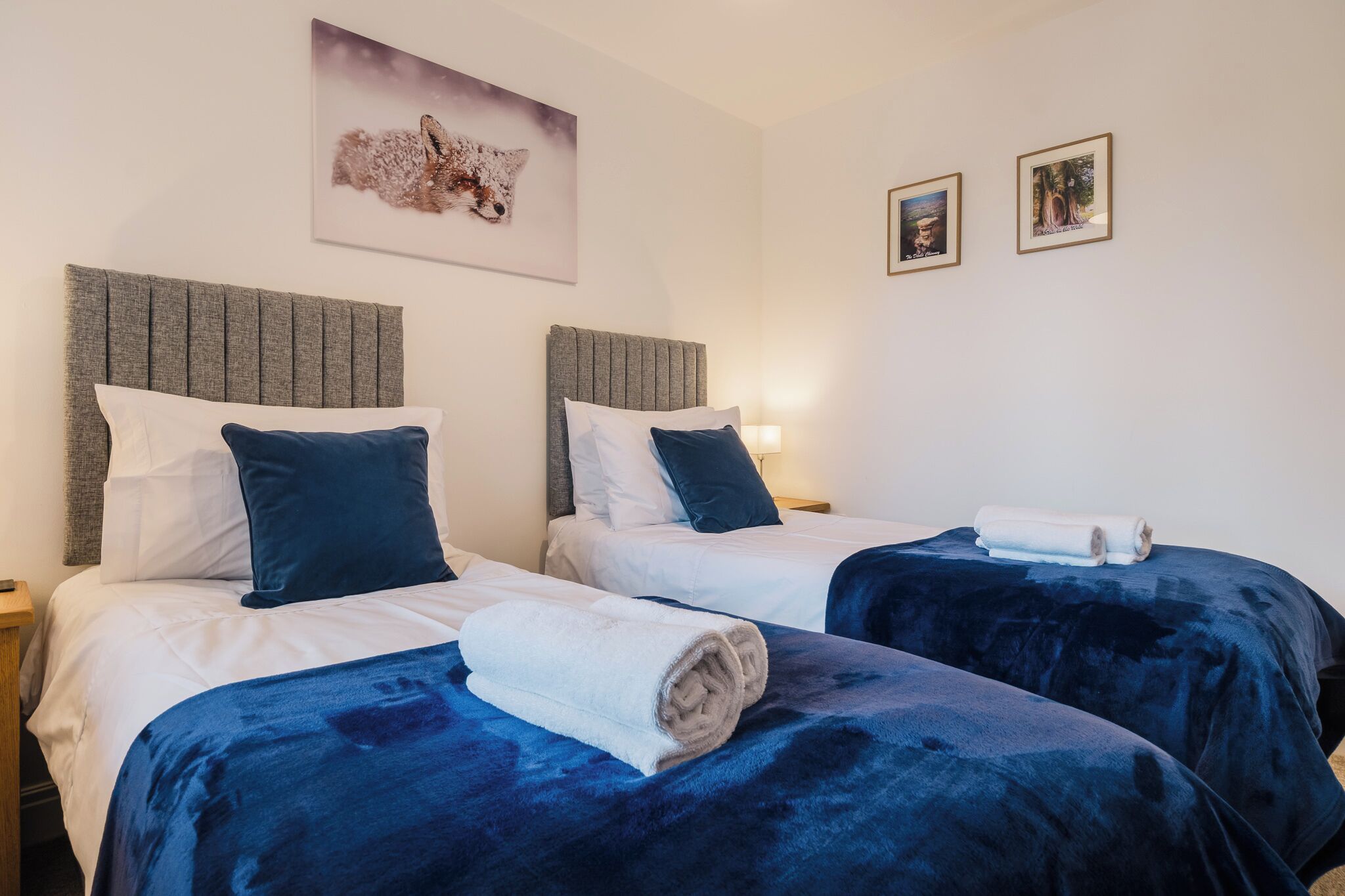 A hotel room with two beds, each with a headboard, white bedding, and blue throw blankets. There are framed pictures on the wall and a rolled towel on the bed.