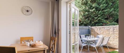 A dining area with a wooden table and chairs, a clock on the wall, and a view of a stone wall and greenery outside.