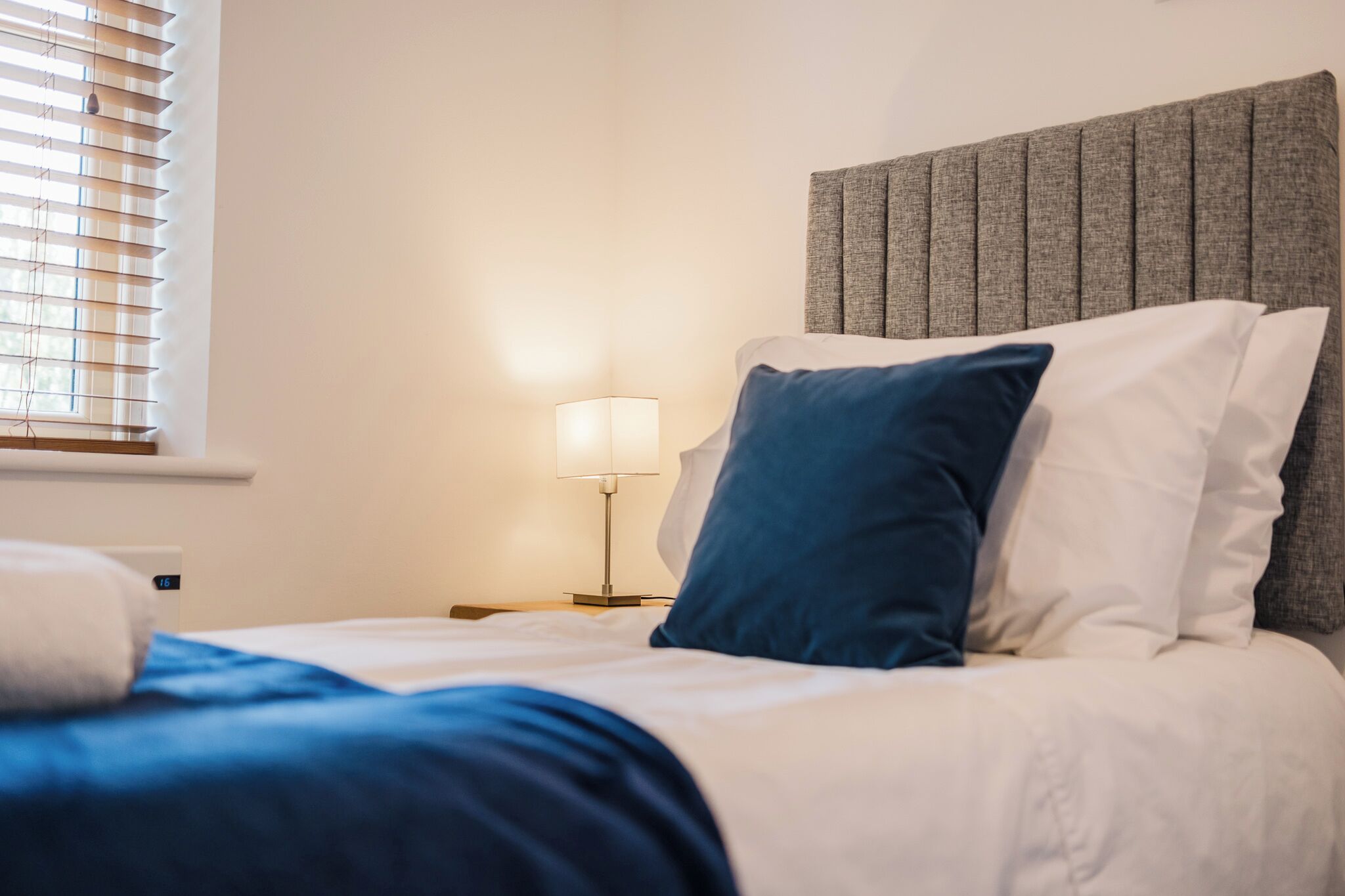 A neatly made bed with white and blue bedding, a grey upholstered headboard, and a bedside table with a lamp.