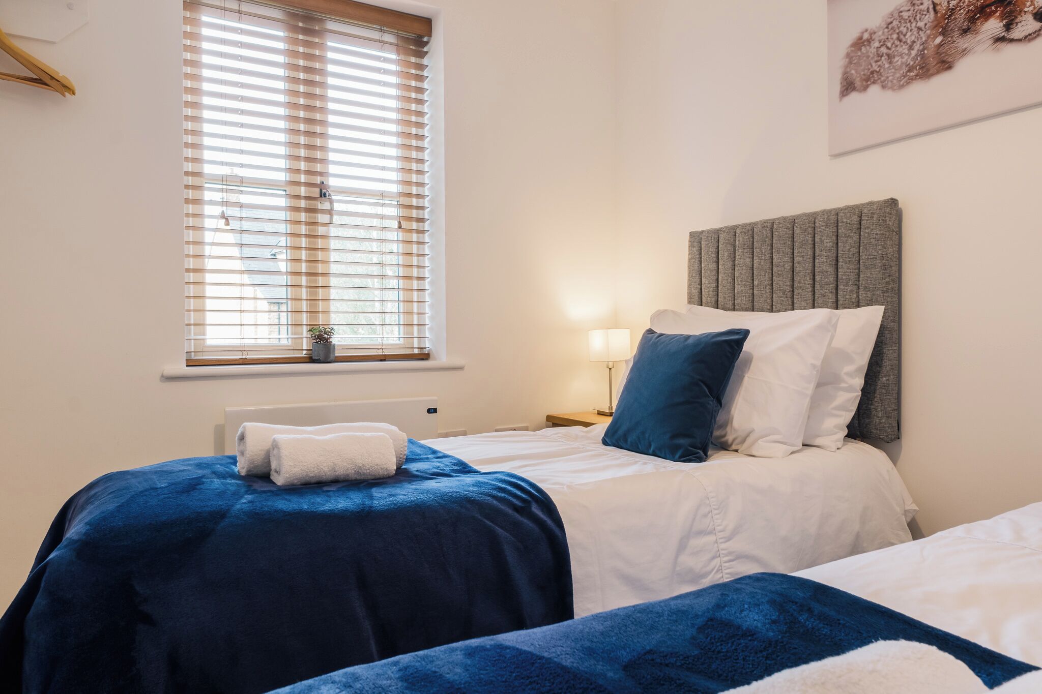 Une chambre d’hôtel avec deux lits, une fenêtre avec des stores, une lampe de chevet et une œuvre d’art accrochée au mur.