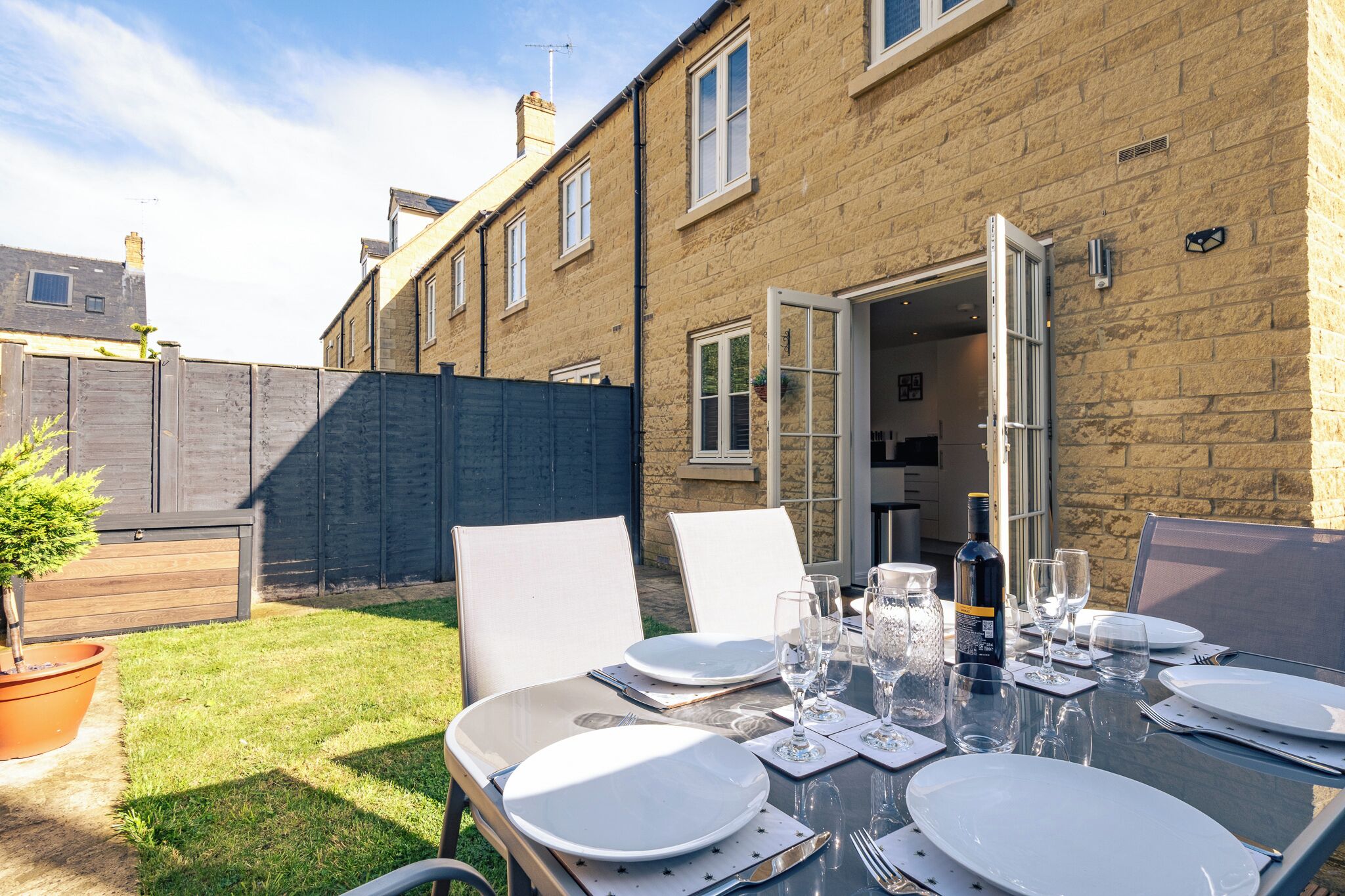 An outdoor dining area with a table set for four, surrounded by chairs, in front of a brick building with an open door.