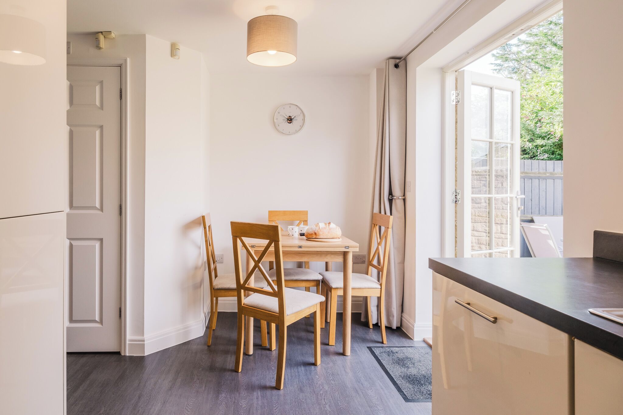 Une cuisine avec une table à manger, quatre chaises, une horloge et une fenêtre avec des rideaux.