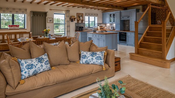 A spacious living area with a brown sofa, wooden beams, a kitchen with blue cabinets, and a wooden staircase. - The Long Barn - Near Oxford (Near Oxford)