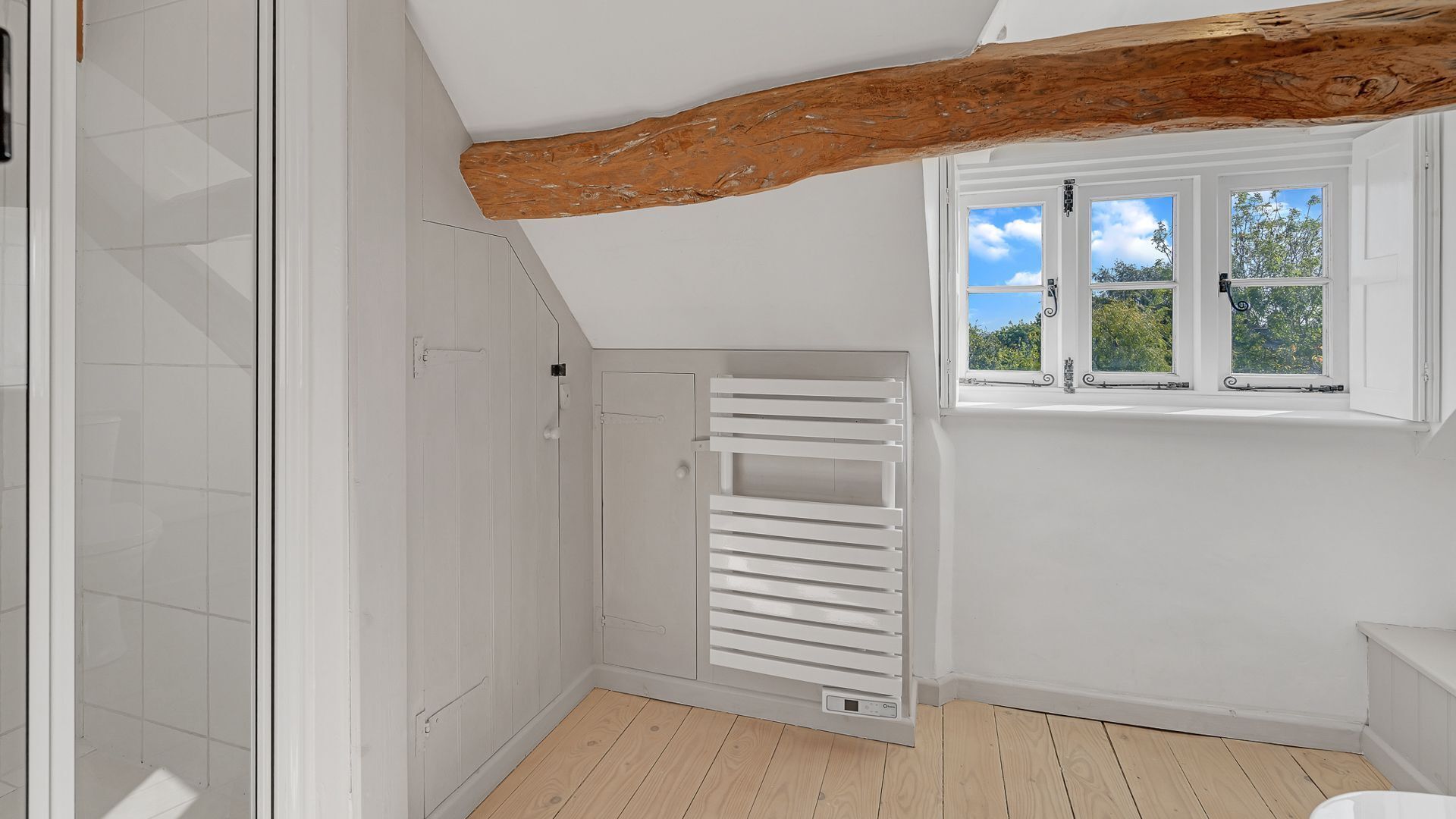 A room with a slatted white wardrobe, a wooden ceiling beam, and a window with a view of greenery.