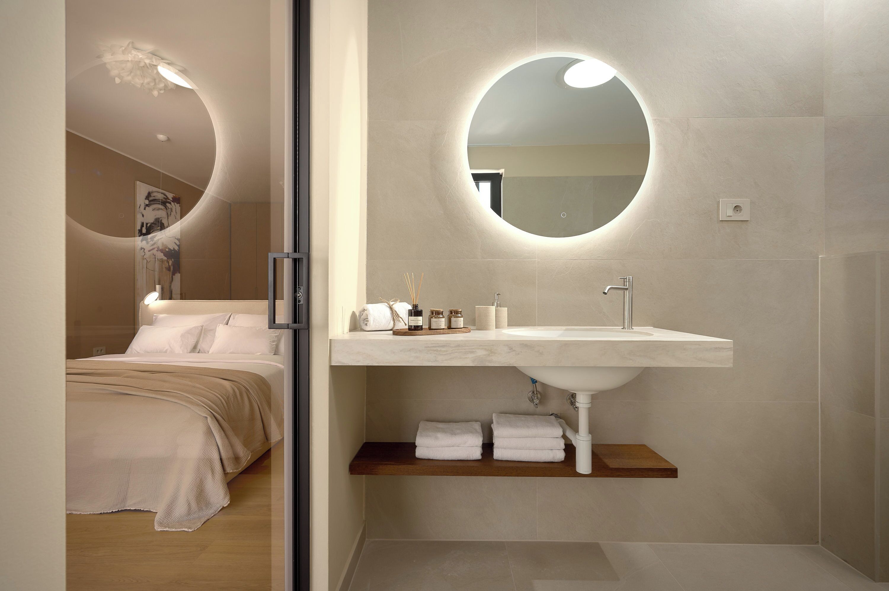 A modern bathroom with a round mirror, a white sink, and a wooden shelf with towels.