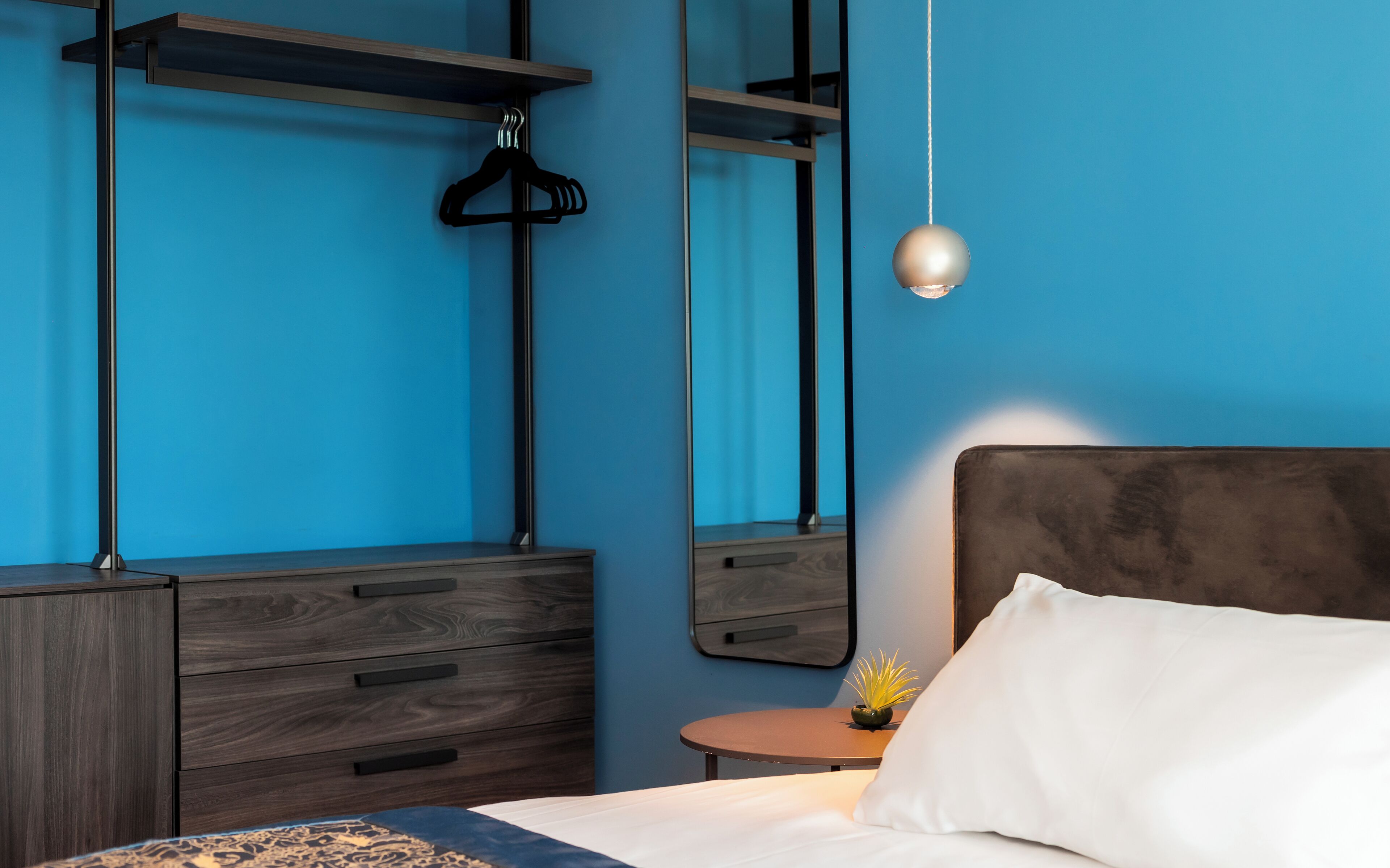 A bedroom with a bed, a dresser, a round table with a plant, and a hanging light fixture.