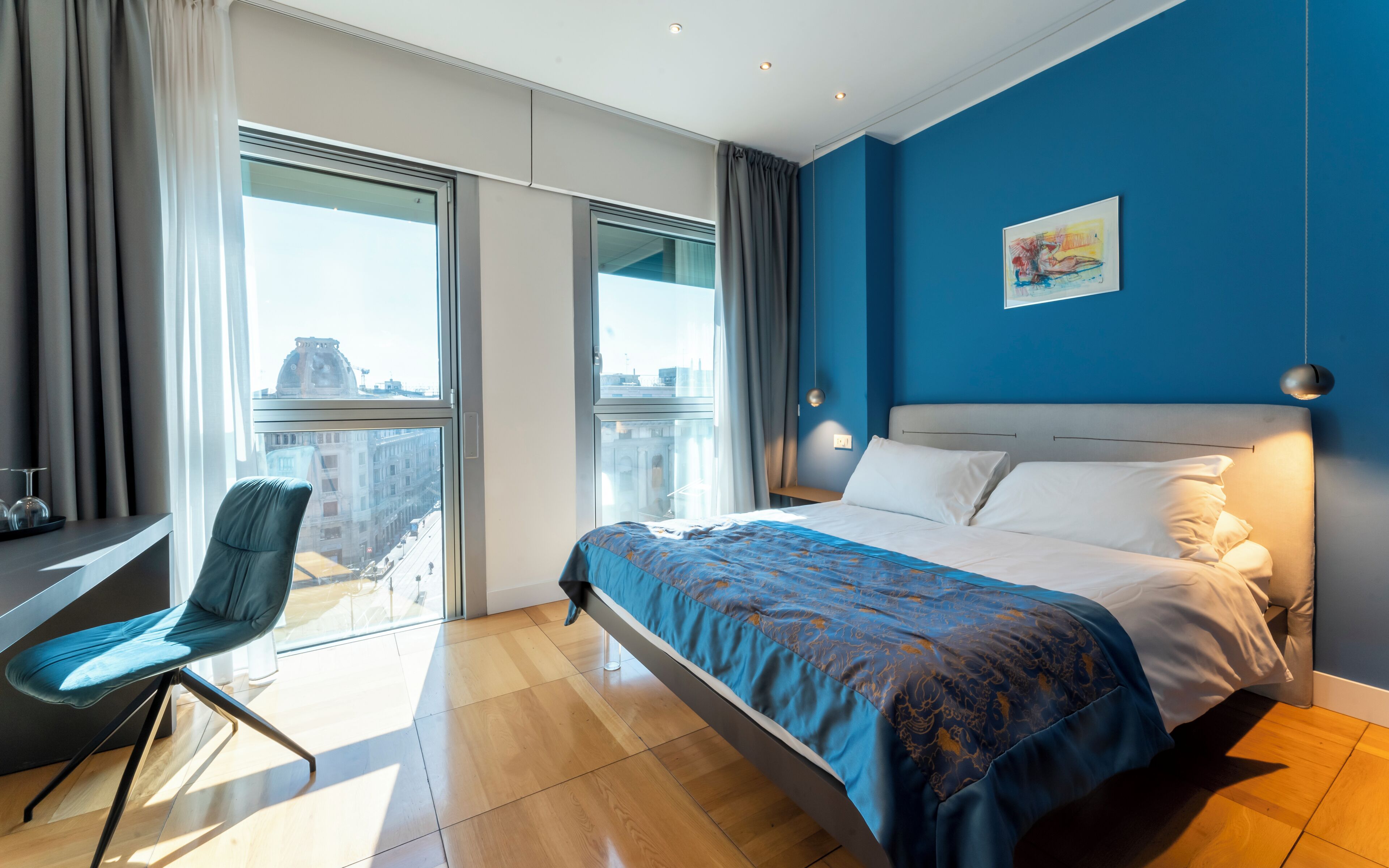 A modern hotel room with a large bed, a desk with a chair, and a view of the city through large windows.