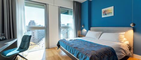 A modern hotel room with a large bed, a desk with a chair, and a view of the city through large windows.