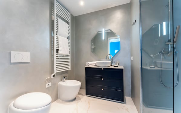 Un baño moderno con inodoro y lavamanos blancos, tocador negro, cabina de ducha con mampara de vidrio y un espejo grande.