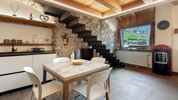 A modern kitchen with a stone wall, wooden staircase, and a dining area with white chairs and a table. - Chez Margot et Sophie – Charm, Stonework and Alpine Views (Torgnon)