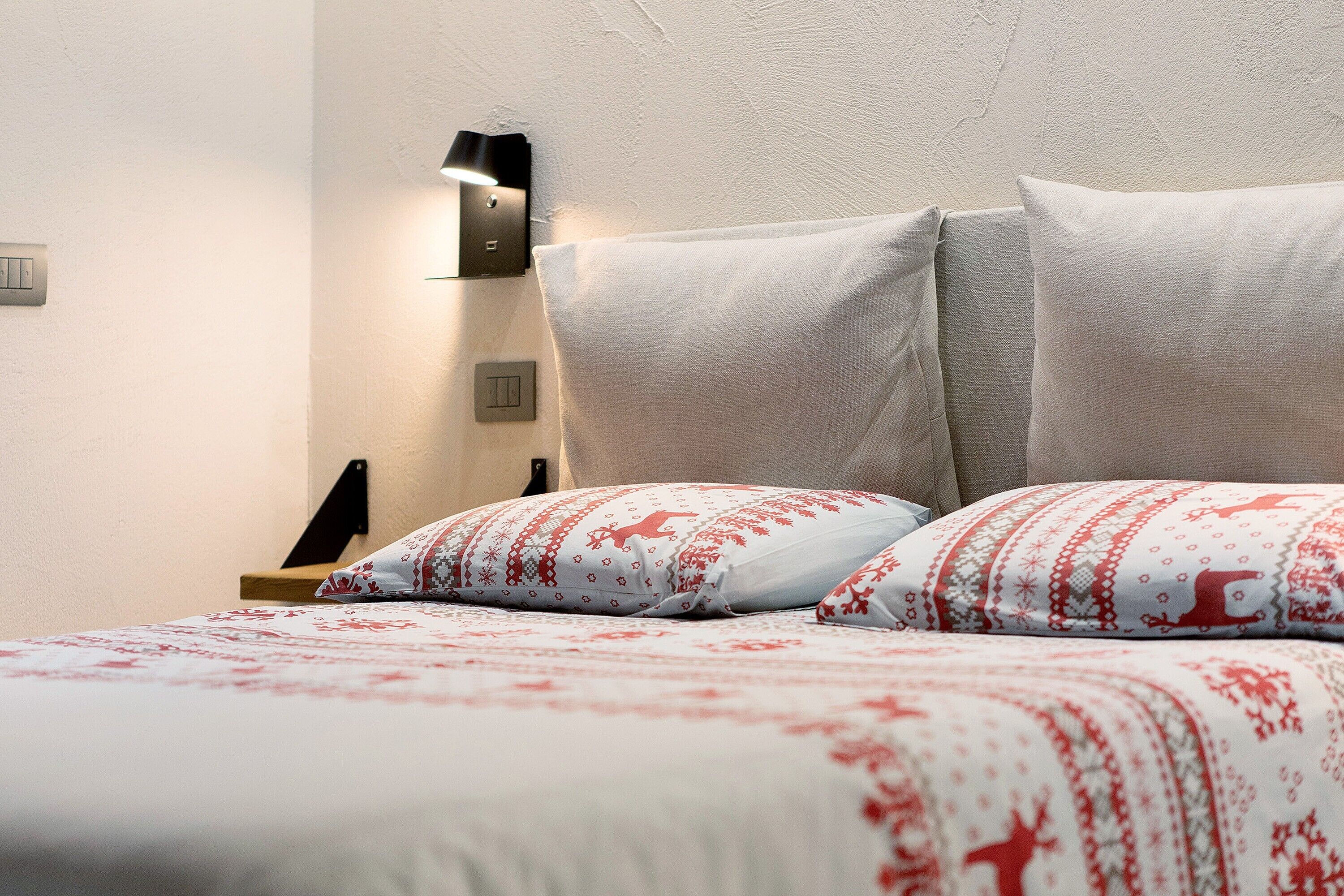 Un lit bien fait, recouvert d’une couvre-lit à motifs, une lampe fixée au mur et un interrupteur près du lit.