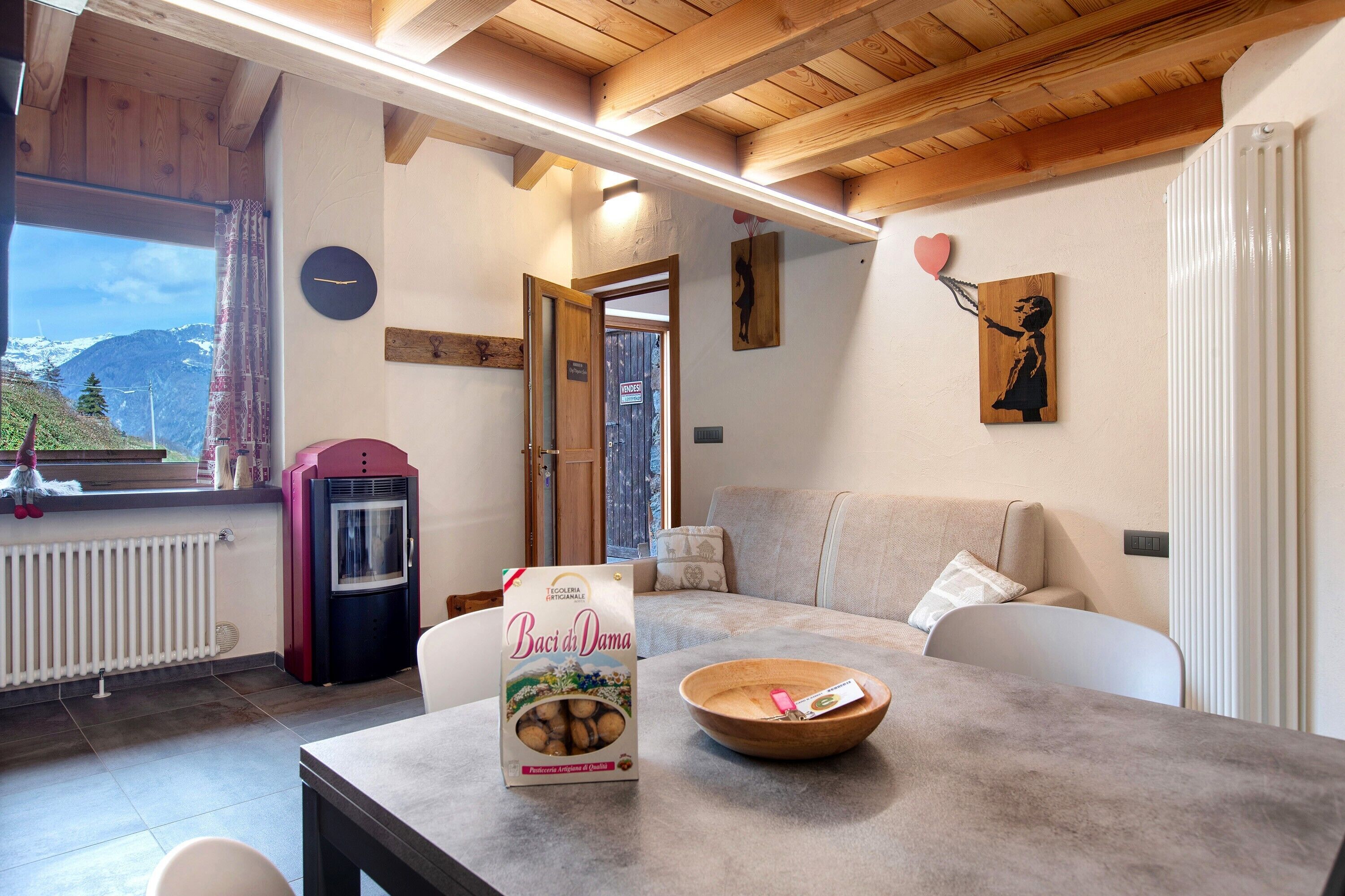 A cozy room with a wooden ceiling, a table with a bowl and a package, a sofa, a radiator, and a window with a view of mountains.